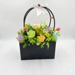Un aranjament floral artificial multicolor într-o geantă neagră, decorat cu o panglică albă și o buburuză decorativă.