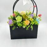 Aranjament floral cu flori decorative multicolore într-o cutie neagră tip geantă, așezat pe o măsuță de cafea."
