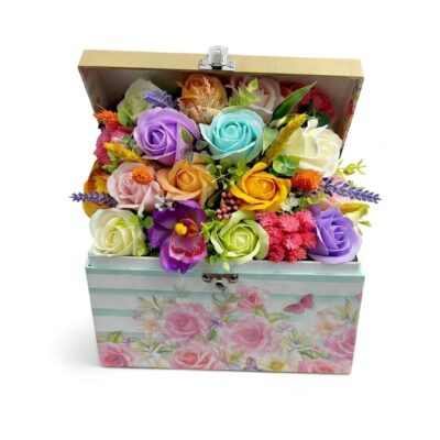 Aranjament floral bogat cu trandafiri și flori din săpun în culori pastelate (mov, bleu, galben, roz), prezentat într-o cutie decorativă cu model floral și capac deschis, pe fundal alb pur."