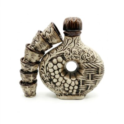 Set tradițional de ceramică artizanala format dintr-o carafă verticală vintage și o stivă de șase pahare mici. Atât carafa, cât și paharele prezintă un model detaliat în relief cu ciorchini de struguri și frunze de viță de vie, cu o glazură mixtă albă și maro, pe fundal alb curat.