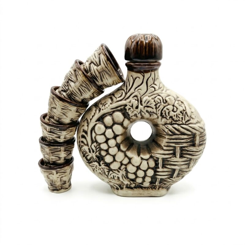 Set tradițional de ceramică artizanala format dintr-o carafă verticală vintage și o stivă de șase pahare mici. Atât carafa, cât și paharele prezintă un model detaliat în relief cu ciorchini de struguri și frunze de viță de vie, cu o glazură mixtă albă și maro, pe fundal alb curat.