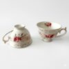Două cești de cafea sau ceai din porțelan alb, cu design vintage elegant, decorate cu motive florale cu trandafiri roz și finisaje aurii pe margini și toartă, prezentate pe fundal alb.