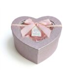 O cutie cadou elegantă, în formă de inimă, acoperită cu sclipici roz și decorată cu o panglică de satin roz cu margine aurie și o fundă centrală. În interior, vizibile prin fereastra transparentă a capacului, sunt aranjate petale de trandafiri din material textil de mătase, nu din flori de săpun. Atașată este o mică etichetă cu urări de bine. Ideal pentru Valentine's Day sau un cadou special pentru o femeie.