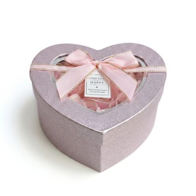 O cutie cadou elegantă, în formă de inimă, acoperită cu sclipici roz și decorată cu o panglică de satin roz cu margine aurie și o fundă centrală. În interior, vizibile prin fereastra transparentă a capacului, sunt aranjate petale de trandafiri din material textil de mătase, nu din flori de săpun. Atașată este o mică etichetă cu urări de bine. Ideal pentru Valentine's Day sau un cadou special pentru o femeie.