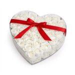 Cutie transparentă în formă de inimă plină cu trandafiri albi artificiali și o fundă roșie de satin pe capac.