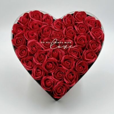 „Cutie elegantă în formă de inimă, plină cu trandafiri roșii din săpun. Pe capacul transparent este inscripționat mesajul 'Do all things with love', prezentată pe un fundal alb, curat.”