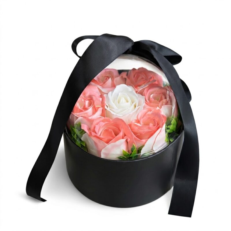 Aranjament floral de lux într-o cutie rotundă neagră, prezentat sub o cupolă transparentă protectoare. Aranjamentul conține un trandafir alb central înconjurat de trandafiri din mătase roz-piersică și frunze verzi decorative. Cutia este accesorizată cu o panglică lată din satin negru, legată elegant într-o fundă mare deasupra cupolei. Ideal ca decor sofisticat sau cadou memorabil.