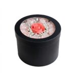 Aranjament floral elegant într-o cutie rotundă de culoare neagră, prevăzută cu capac cu fereastră transparentă. În centru se află un trandafir de culoare coral (somon), înconjurat de trandafiri roz pal realizați din material textil de mătase și mici decorațiuni albe tip bobițe. Produs premium pentru cadouri, decorat cu atenție la detalii.