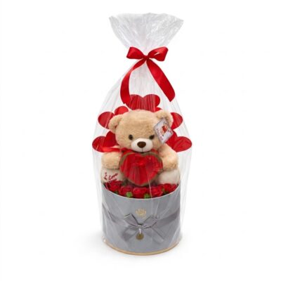 Un set cadou romantic și complet pentru Ziua Îndrăgostiților (Valentines Day), ambalat elegant în celofan transparent cu o fundă roșie și decorat cu decupaje în formă de inimă roșie. În interior se află un ursuleț de pluș bej care ține o cutie de ciocolată în formă de inimă roșie, așezat pe o bază de trandafiri roșii din săpun sau mătase. Ursulețul are o etichetă vizibilă "Hug me" și pe piciorul său este brodat textul "I Love You". Totul este prezentat într-o cutie cilindrică gri decorată, gata de oferit.