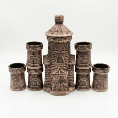 Set rustic din ceramică pentru băuturi, format dintr-o sticlă în formă de turn de castel și pahare asortate, prezentat pe un birou din lemn alături de o carte veche și o lumânare aprinsă, sugerând o atmosferă medievală.
