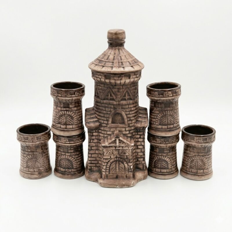 Set rustic din ceramică pentru băuturi, format dintr-o sticlă în formă de turn de castel și pahare asortate, prezentat pe un birou din lemn alături de o carte veche și o lumânare aprinsă, sugerând o atmosferă medievală.