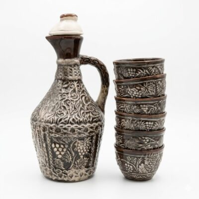 Set tradițional din ceramică pentru vin, format dintr-un ulcior cu capac și șase cești suprapuse. Piesele sunt maro cu detalii gri și au motive în relief cu struguri și frunze de viță de vie, prezentate pe fundal alb.
