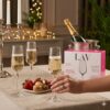 Pahare de șampanie LAV Lal pline, servite la o masă festivă elegantă cu macarons, căpșuni și lumânări, alături de cutia produsului.
