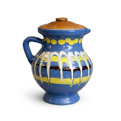 O fotografie de studio pătrată a unui ulcior ceramic albastru strălucitor, lucrat manual, cu un capac de lut de culoare maro-auriu. Ulciorul are un gât cu buline galbene și un corp central decorat cu benzi orizontale galbene și negre, peste care coboară un model distinctiv de picurare vertical alb și galben. Are un mâner pe partea stângă și o bază profilată. Fundalul este alb pur.