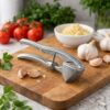 Presă de usturoi din metal așezată pe un tocător de lemn, alături de căței de usturoi și roșii.